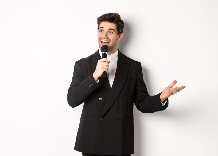 Portrait of handsome man in black suit singing a song, holding microphone and giving speech, standing against white background.