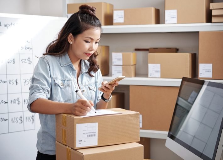 Asian woman looking address in her mobile phone and writing it on parcel at post office